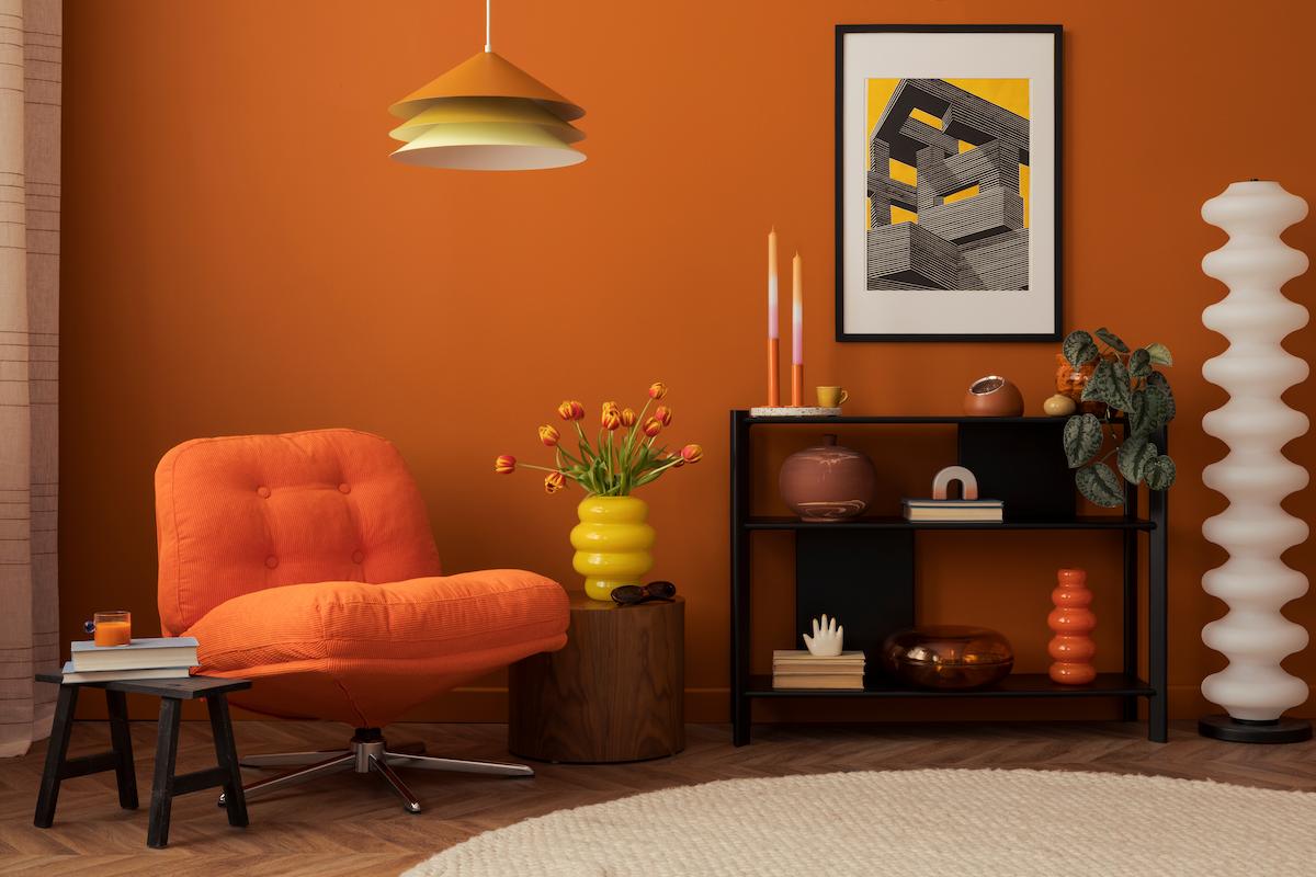 Salon aux murs orange, avec un fauteuil orange, des objets décos aux formes organiques, une lampe en métal et une table d'appoint en bois foncé